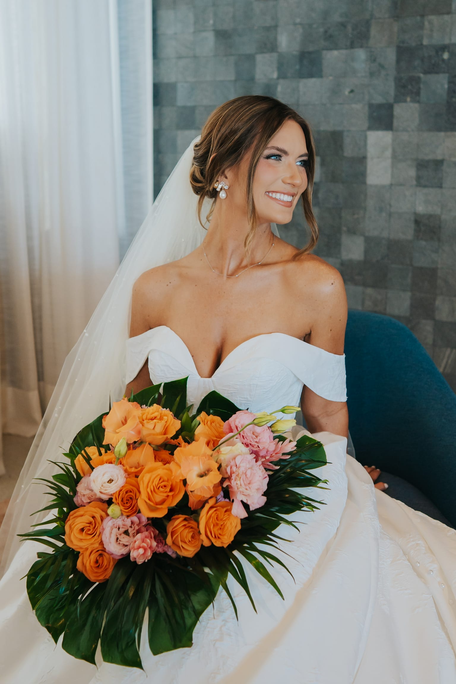 Smiling bride in a Madison and Blake off-shoulder white dress holds orange-pink bouquet at wedding.