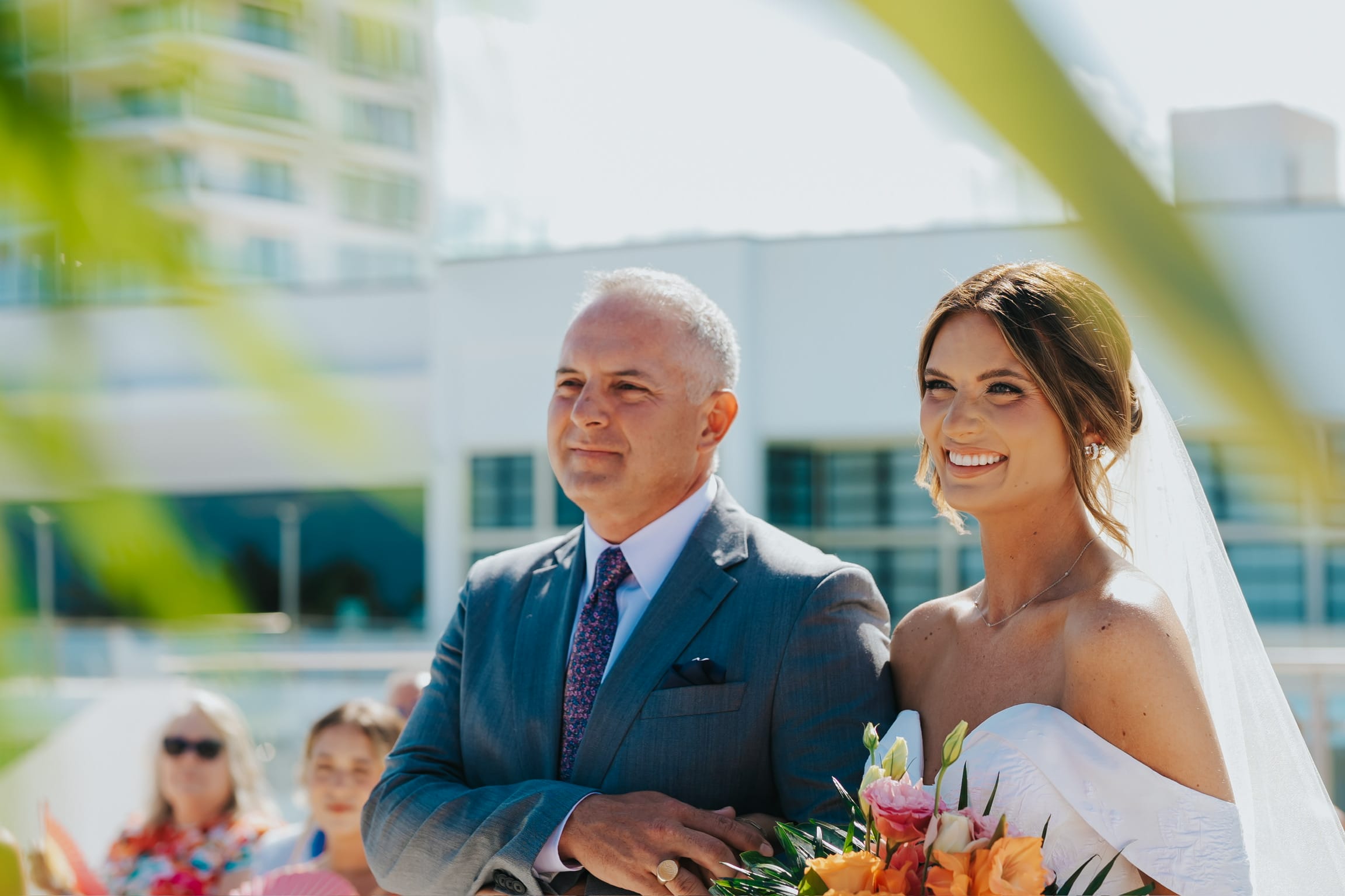 Bride holding Madison and Blake bouquet stands beside man in suit at outdoor destination wedding ceremony.