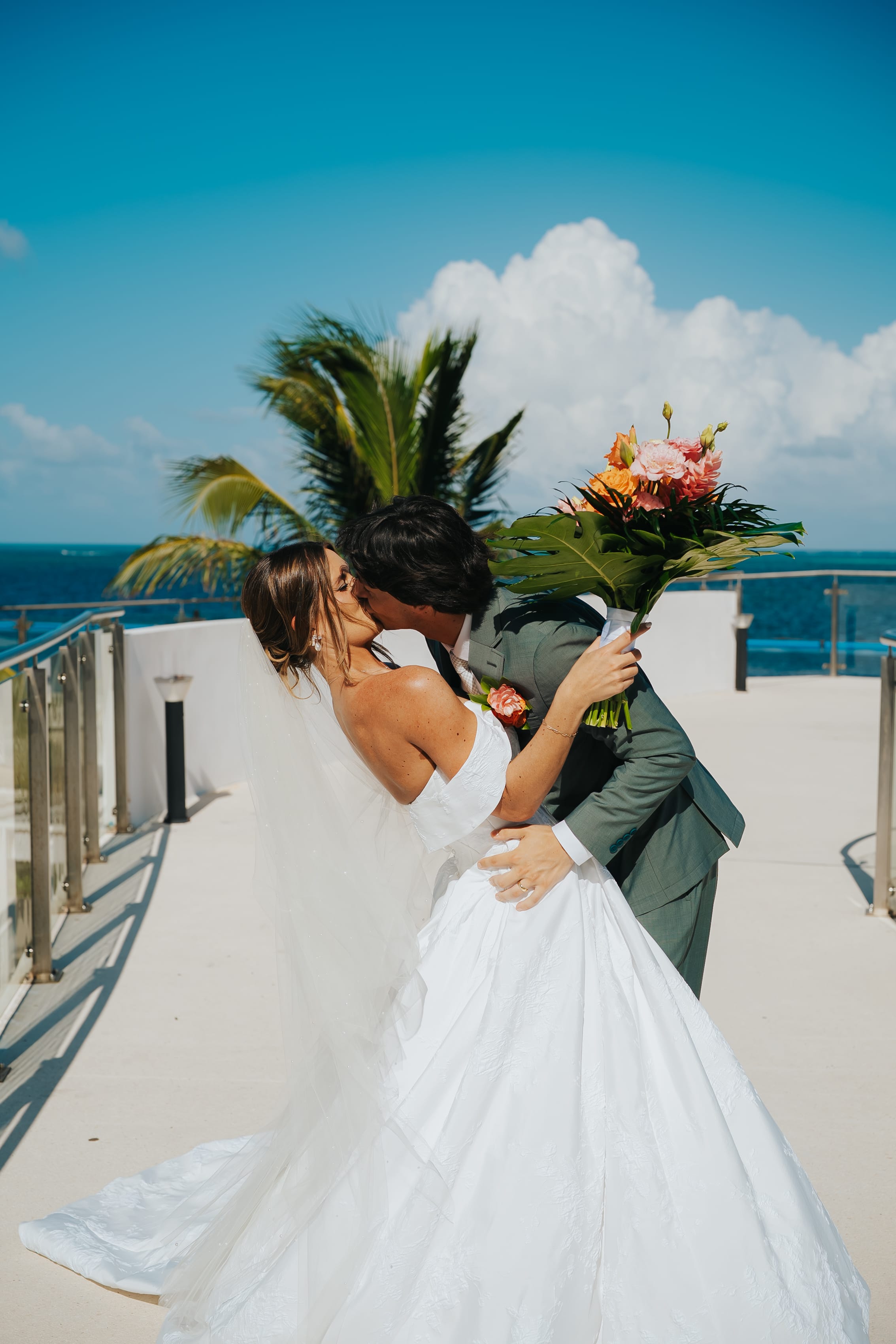 Madison and Blake share a kiss at their destination wedding by the ocean, with palm trees and blue sky.