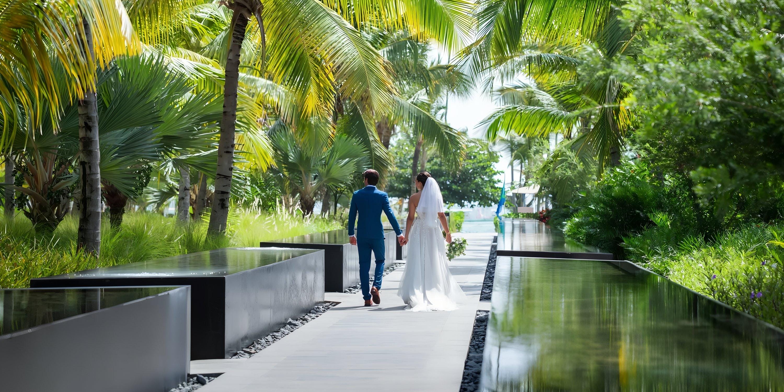 Bride and groom stroll hand in hand along a palm-lined path at their destination wedding in Costa Bavaro.